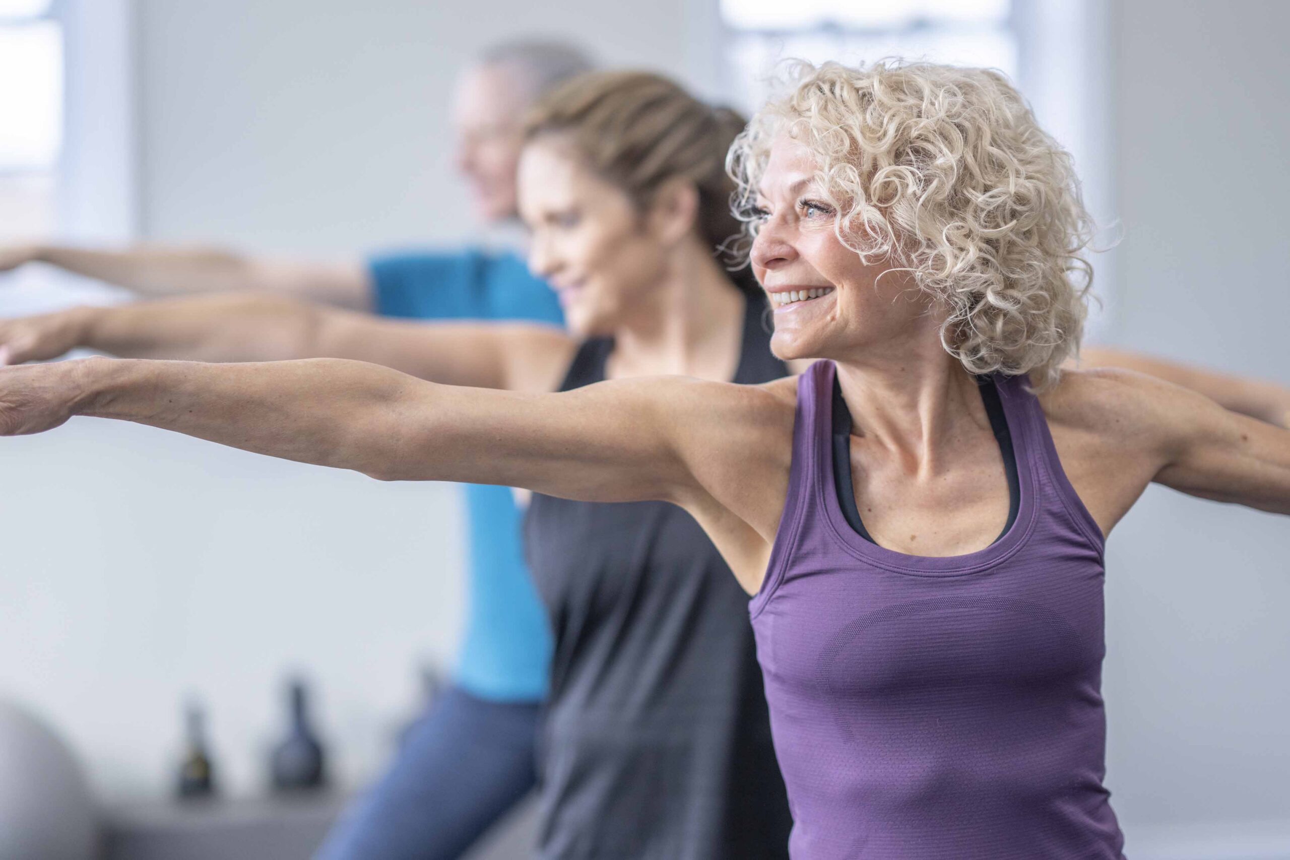 A Small Group Of Three Mature Women Stand Spread Apart On Individual Yoga Mats As They Participate In A Class Together.  They Are Each Dressed Comfortably In Athletic Wear And Are Holding A Warrior Pose.